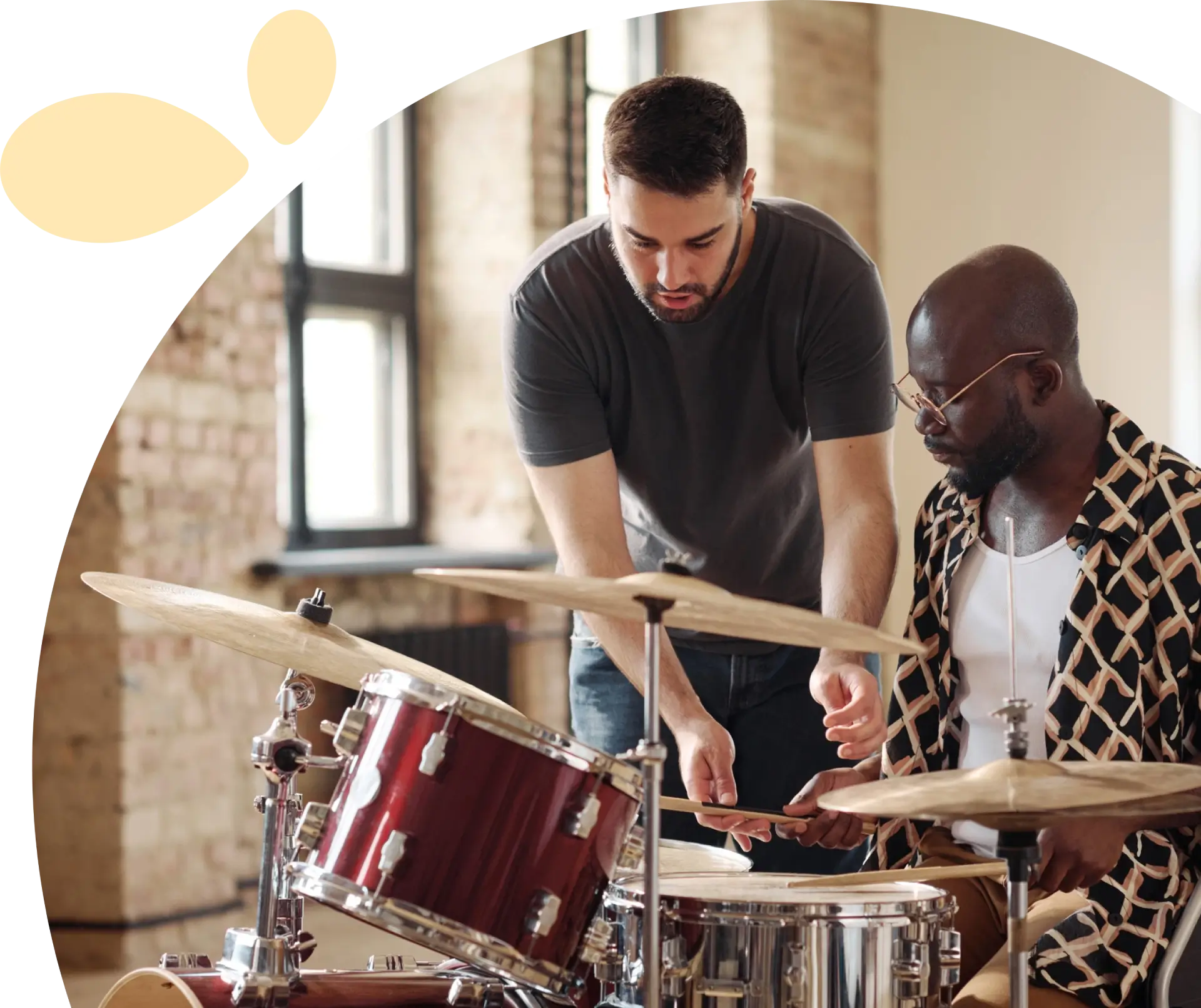 a22Frame 1618873785 (1) (1) Two men in a room with large windows; one man plays drums while the other instructs, pointing at the drum kit.