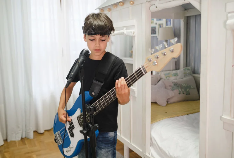 A young boy wearing a headband and black shirt plays a blue electric guitar in front of a microphone. Bedroom background with a bed.