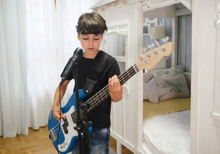 A young boy wearing a headband and black shirt plays a blue electric guitar in front of a microphone. Bedroom background with a bed.